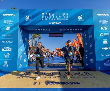 Photographie de deux coureurs franchissant la ligne d’arrivée du Marathon des Alpes-Maritimes Nice-Cannes. Au premier plan, une femme et un homme, souriants, se tiennent la main en levant les bras dans un geste de joie. Ils portent des tenues de sport, des dossards et des chaussures de course. Ils passent sous une grande arche bleue décorée des logos des partenaires de l’événement. Au sol, le nom du marathon est inscrit en grandes lettres bleues. À l’arrière-plan, d’autres coureurs poursuivent leur course sous un ciel bleu ensoleillé, bordé de palmiers. L’image exprime la joie, l’effort sportif et le plaisir de franchir ensemble la ligne d’arrivée.